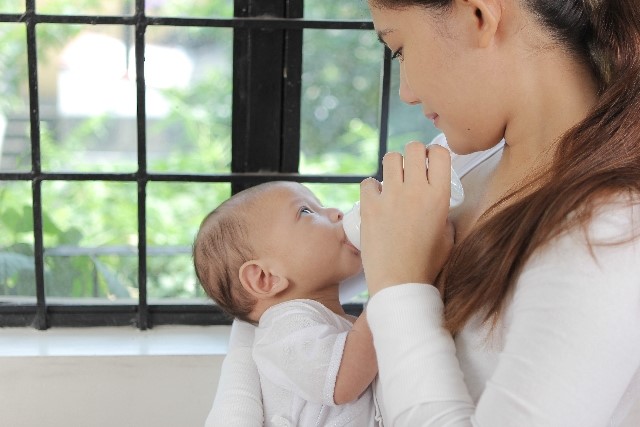 窓辺で女性が赤ちゃんにミルクを与えている写真